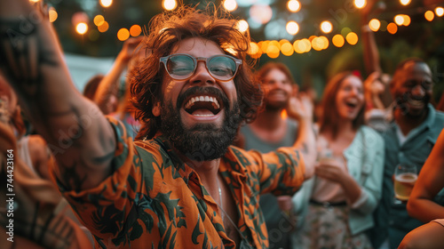 A bearded man in glasses is captured in a moment of pure joy, surrounded by friends at an outdoor party with festive lights.
