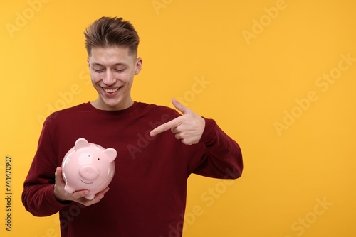 Happy man pointing at piggy bank on yellow background. Space for text