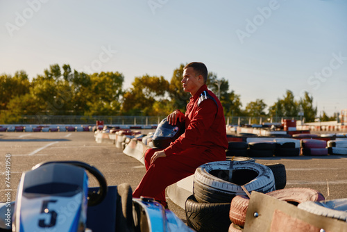 Male racer in protective overalls holding helmet on go-kart track outdoors