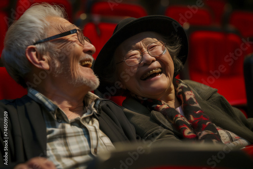 Happy chinese or japanese old couple man and woman having fun at the cinema watching alone a movie sitting on red seats laughing a lot. Enjoying theatre entertainment mature people from east photo