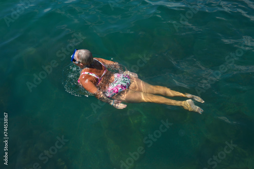 Aged woman is swimming with goggles in green sea water.