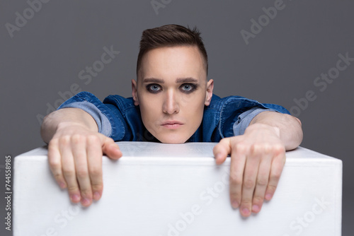 Portrait of a young man with rock musician makeup sitting at a white cube