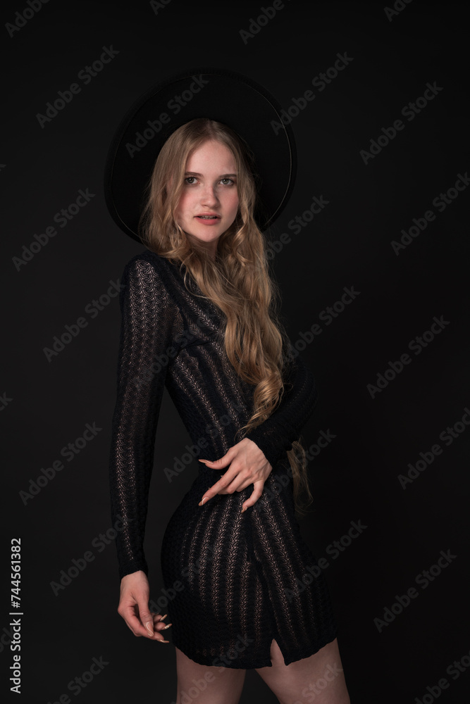Slim woman striking graceful pose with one hand on waist. Black background accentuates her beauty, while her black short knitted translucent dress and wide brimmed felt hat add touch of elegance Slim woman striking graceful pose with one hand on waist. Black background accentuates her beauty, while her black short knitted translucent dress and wide brimmed felt hat add touch of elegance