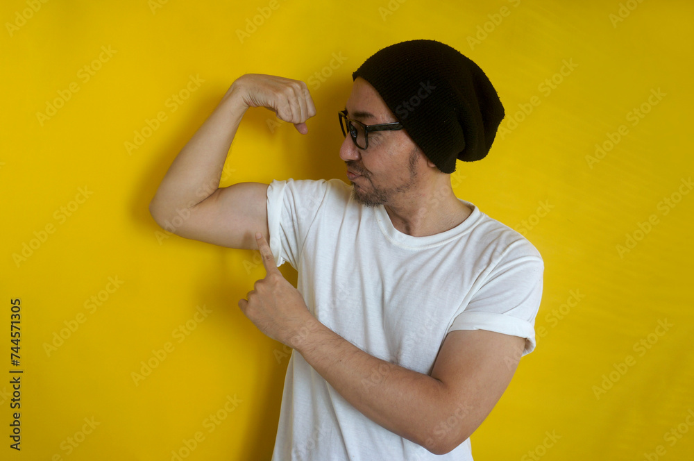 Asian hipster man wear beanie hat and eyeglasses, showing strong hand muscle biceps over yellow background Asian hipster man wear beanie hat and eyeglasses, showing strong hand muscle biceps over yellow background