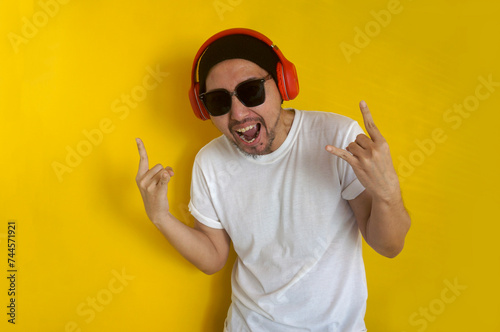 Excited man with sunglasses and beanie hat listening to music using headphones shouting with crazy expression doing rock symbol with hands up