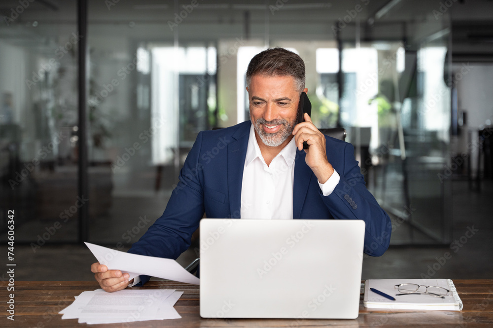 Middle aged Latin or Indian businessman having call on smartphone with business partners or clients. Smiling mature Hispanic man sitting at table talking by mobile cellphone and check data in office. Middle aged Latin or Indian businessman having call on smartphone with business partners or clients. Smiling mature Hispanic man sitting at table talking by mobile cellphone and check data in office.