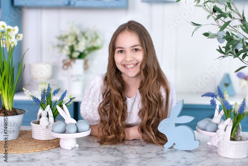 Child Girl in dress with Easter eggs at home. Portrait of happy little girl with bouquet flowers in kitchen. Smiling Girl is preparing for Easter. Little Girl hunts and picks up easter eggs from table photo