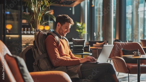 Smiling Man with Backpack Working on Laptop in Stylish Cafe Interior © Anna