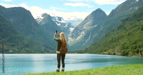 Woman, lake and cellphone for pictures, mountain and norway with backpack, view and sightseeing. Technology, exploring and nature for vacation, adventure and travelling in outdoor for tourism photo