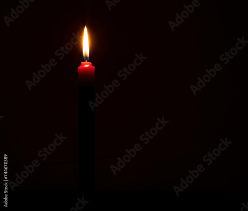 Single burning candle flame or light glowing on a small red candle on black or dark background on table in church for Christmas, funeral or memorial service with copy space.