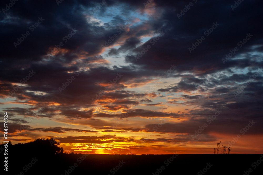 Sunset in the summer field. The clouds in the sky are lit by the setting sun on the horizon. The blue sky changes color in the rays of the sun at sunset. Clouds in the sky above the horizon.