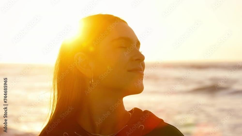 Head and shoulders shot of casually dressed young woman sitting on sand watching beautiful sunrise morning over beach and sea in South Africa with flaring sun  - shot in slow motion