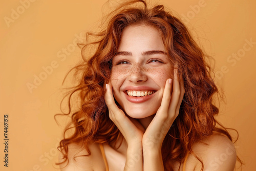Joyful young woman with freckles smiling with hands on cheeks on a beige background.
