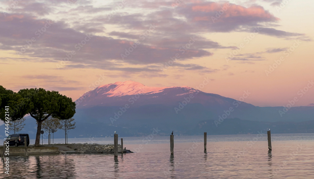 Picturesque natural landscape with stunning sunset over serene lake framed by majestic snow-capped mountain peak in background. Lake Garda, Sirmione, Italy, Alps. Mobile photo.