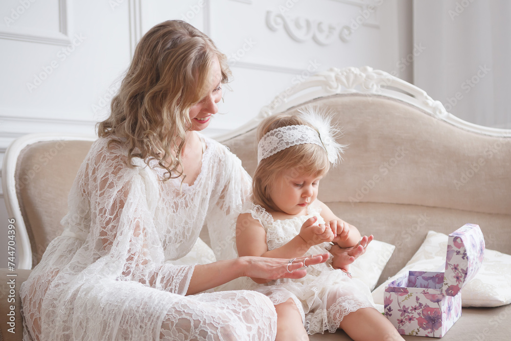 A mother and a daughter in lace dresses are sitting on a sofa in white interior living room. A little girl putting many rings on her mother's fingers. A mother and a daughter in lace dresses are sitting on a sofa in white interior living room. A little girl putting many rings on her mother's fingers.