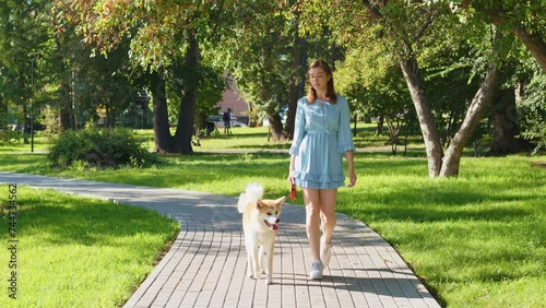 Beautiful girl in dress walks dog on leash in urban park. Young woman going with Akita Inu along paved path surrounded by grass, trees. Caucasian lady with pet spend time on city street in summer photo