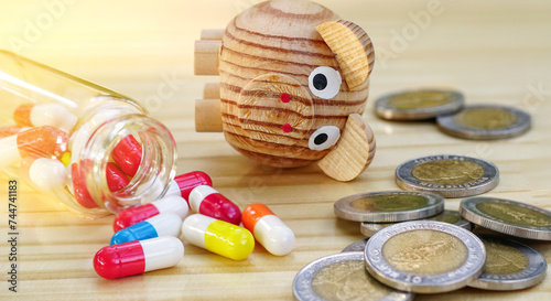 Miniature wooden piggy bank falls overturned with heap of money coins and colorful medicine pills on wood table, Money debt problem in medication concept.Crisis of drug high cost in disease treatment.