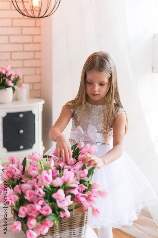 Little girl is standing in the bedroom. Large bouquet of tulip flowers. Princess in a white dress with a bouquet of pink flowers in the room. Little girl is standing in the bedroom. Large bouquet of tulip flowers. Princess in a white dress with a bouquet of pink flowers in the room.