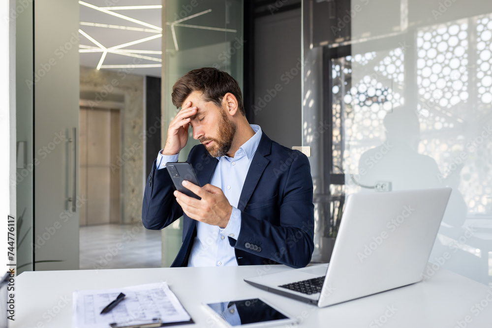 Shocked young man businessman sitting in the office on a chair and holding his head with his hand, holding the phone, upset by the news he received Shocked young man businessman sitting in the office on a chair and holding his head with his hand, holding the phone, upset by the news he received