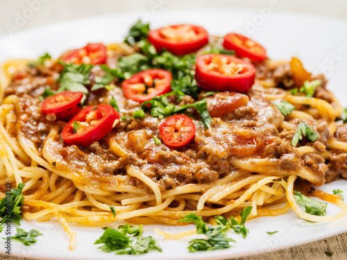 Spaghetti Bolognese dish with pasta, red sauce, minced beef, red peppers and green fresh herbs on white plate and light color table cloth. High quality products in a tasty Italian style dish.