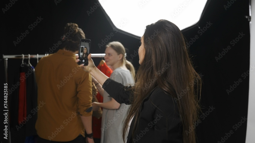 Backstage of model and professional team in the studio. Appealing brunette model taking selfie on smartphone, photographer and girl at the back.
