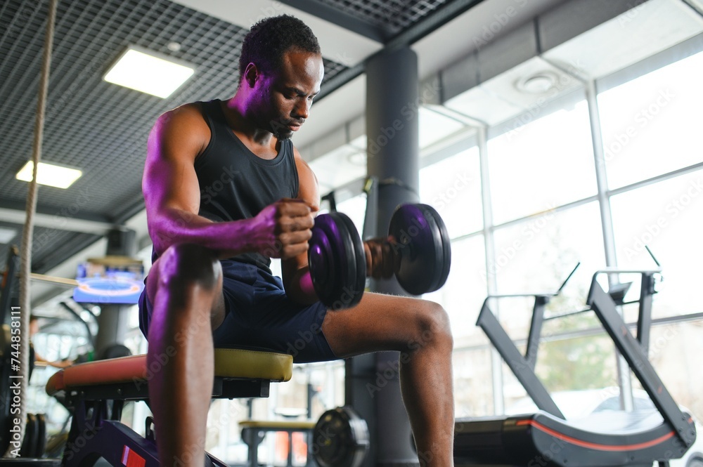 Young African American man sitting and lifting a dumbbell close to the rack at gym