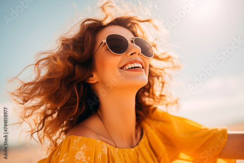 A joyful woman in a yellow dress with sunglasses  her hair playfully tousled by the wind  laughing against a clear sky.