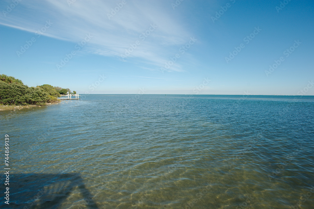 Obraz premium View out from Bay Vista Park pier east towards Tampa Bay on a sunny afternoon in St. Petersburg, FL. Sunlight and shade of pier in blue water and peaceful. Green mangroves and boat dock jetty.