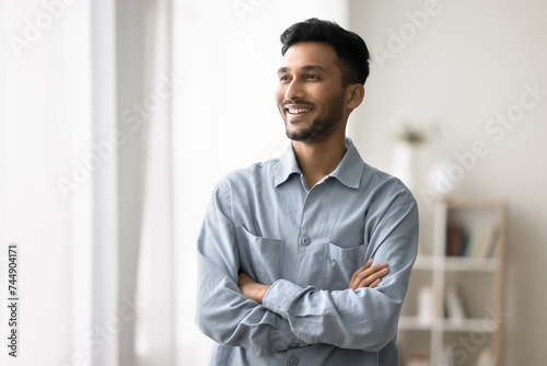 Young Indian man spend time at home, standing with arms crossed on chest smile, look away, enjoy day-off at own or rented apartment, rest, relish pleasant thoughts, good life opportunity and leisure photo