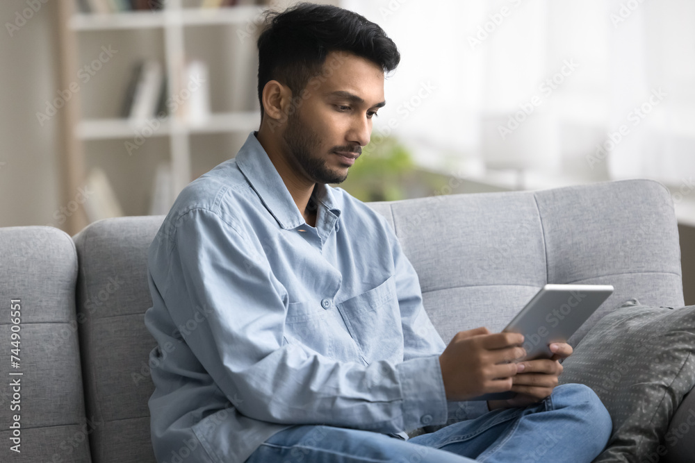 Serious Indian guy read news using digital tablet resting on couch, websurfing internet spend free time on weekend search information, learn new application, program or application. Modern tech usage Serious Indian guy read news using digital tablet resting on couch, websurfing internet spend free time on weekend search information, learn new application, program or application. Modern tech usage