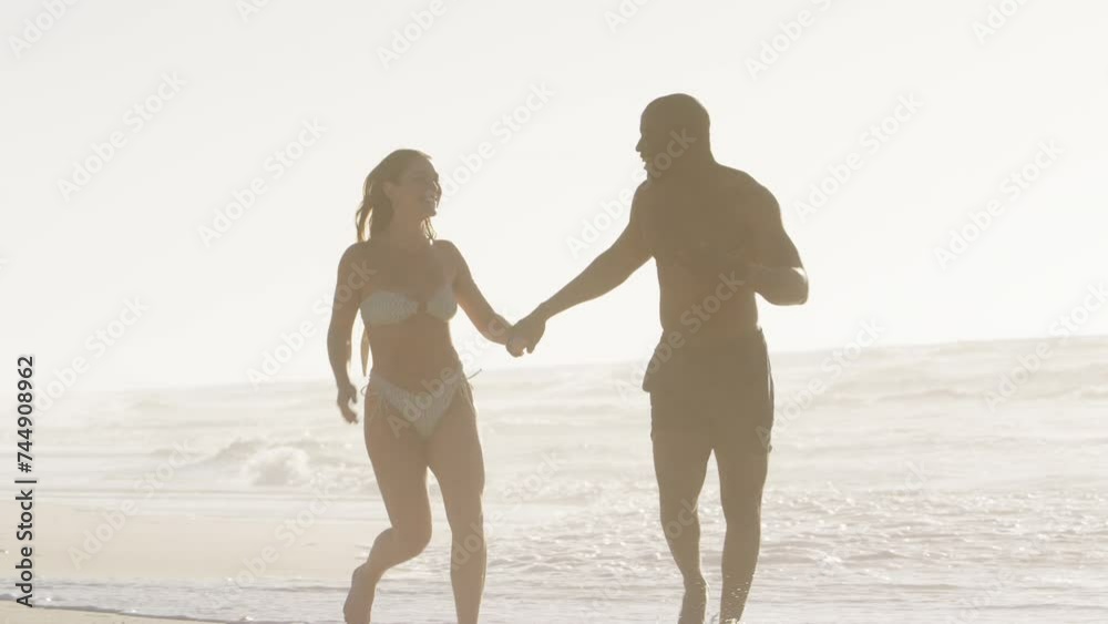Three quarter length shot of loving young couple in swimwear holding hands and running along beach in South Africa - shot in slow motion Three quarter length shot of loving young couple in swimwear holding hands and running along beach in South Africa - shot in slow motion