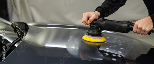 A mechanic polishes the surface of the hood of a gray car. 