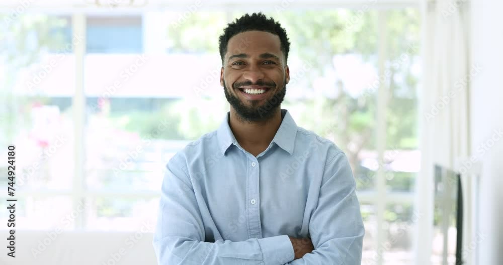 Confident handsome young African man in office shirt looking at camera with arms crossed, posing for head shot portrait indoors with large window in background, getting happy, cheerful, smiling Confident handsome young African man in office shirt looking at camera with arms crossed, posing for head shot portrait indoors with large window in background, getting happy, cheerful, smiling