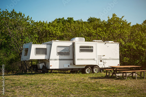 Travel trailer stands in meadow near the forest at dawn. The travel trailer is parked in the trailer parking. Wooden table and picnic benches.