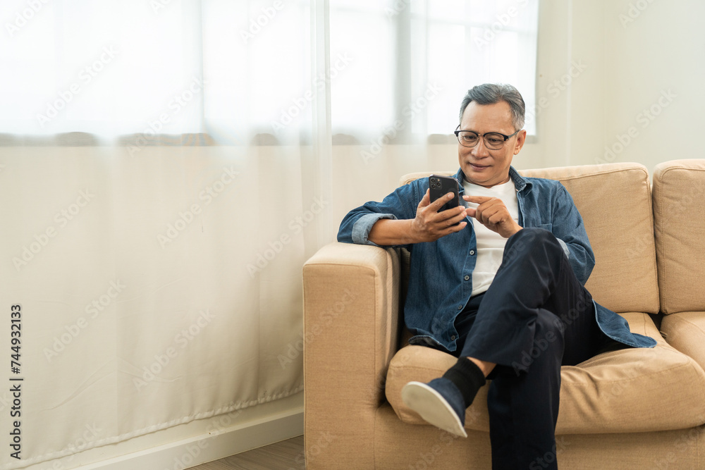 Handsome asian mature old man using smartphone on sofa in living room at home. Happy Portrait of cheerful smiling senior asian man holding cell phone. Mature People and lifestyle Handsome asian mature old man using smartphone on sofa in living room at home. Happy Portrait of cheerful smiling senior asian man holding cell phone. Mature People and lifestyle