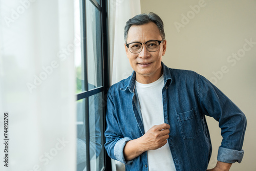 Handsome asian mature old man standing near windows with sunlight in the morning. Happy Portrait of cheerful smiling senior asian man pose looking at camera. Mature People and lifestyle photo
