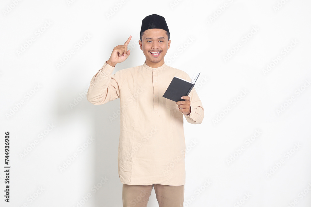 Portrait of attractive Asian muslim man in koko shirt with peci reading a book, telling that he has an idea while pointing finger and pen. Isolated image on white background Portrait of attractive Asian muslim man in koko shirt with peci reading a book, telling that he has an idea while pointing finger and pen. Isolated image on white background