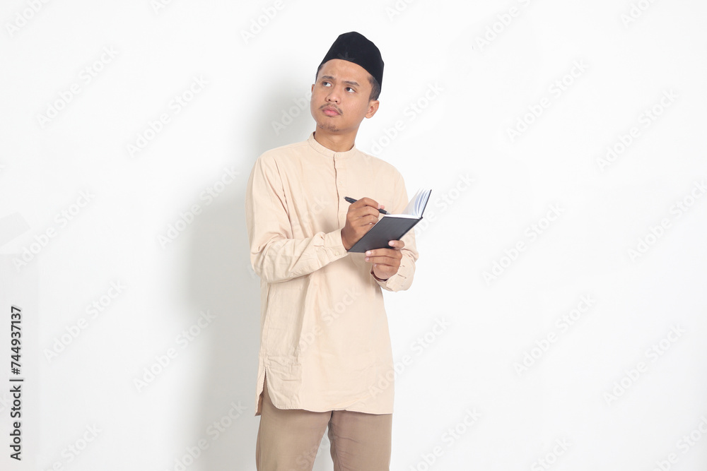 Portrait of confused Asian muslim man in koko shirt with peci reading a book, thinking about an idea with hand holding pen on chin, looking away. Isolated image on white background Portrait of confused Asian muslim man in koko shirt with peci reading a book, thinking about an idea with hand holding pen on chin, looking away. Isolated image on white background