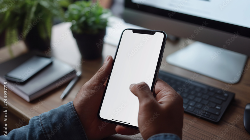 Close-up of a professional's hand holding a smartphone with a blank screen, with a laptop, coffee cup, and eyeglasses in the background on a desk.