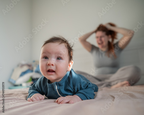 A three-month-old boy lies on his stomach on the bed and his mother sits behind him and cries. Postpartum depression.