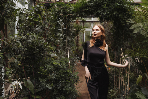 A woman with red hair, dressed in a black dress, walks through a lush green greenhouse. Concept for promoting cosmetic products or perfume for an elegant lifestyle.