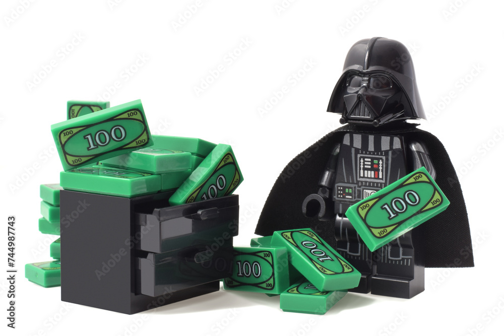 Naklejka premium Lego minifigure of Darth Vader or Star Wars male is counting his money green banknotes isolated on white. Editorial illustrative image of finance and banking.