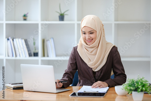 young pretty modern muslim woman in hijab working on laptop in office room, education online, remote work freelancer, using smartphone © Treerat