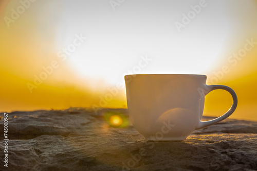 A white cup of hot espresso coffee mugs on the rocks and nature view of the sun background in the morning with sunlight