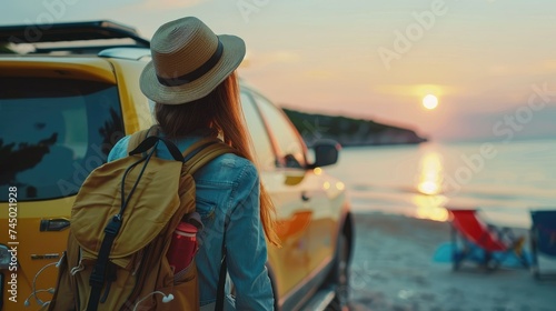 Young woman with a backpack standing on the beach and looking at the sunset. She is wearing a hat and a denim shirt.