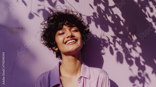 A joyful individual with short hair smiling against a purple wall with leaf shadows.