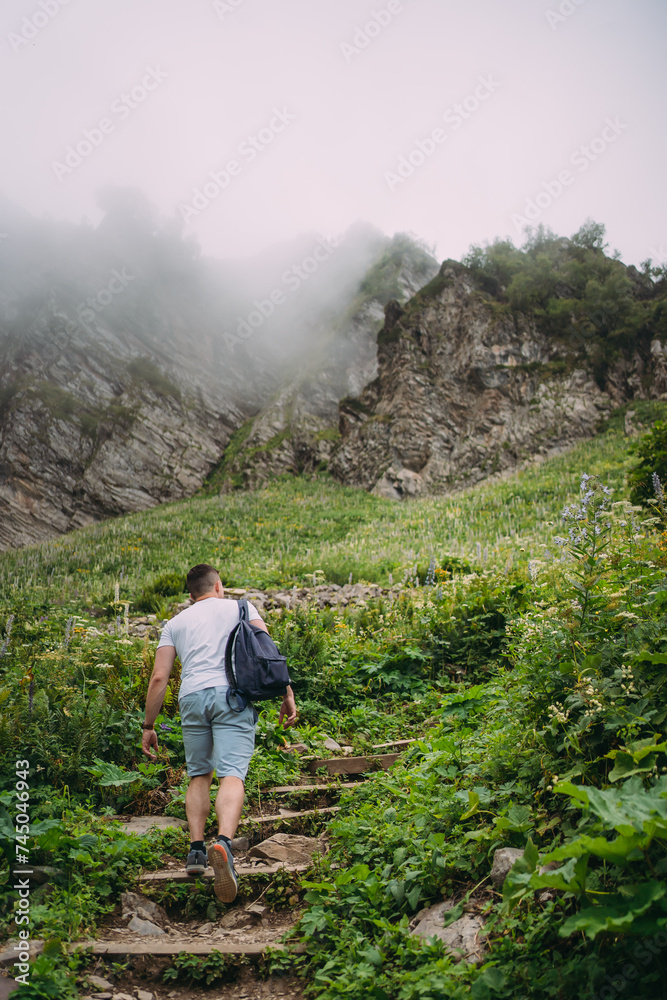 Naklejka premium A man climbs the stairs in the mountains. Hiking trails high in the mountains in summer. The man is wearing a white T-shirt, blue shorts and a blue backpack. A cloud enveloped the mountain