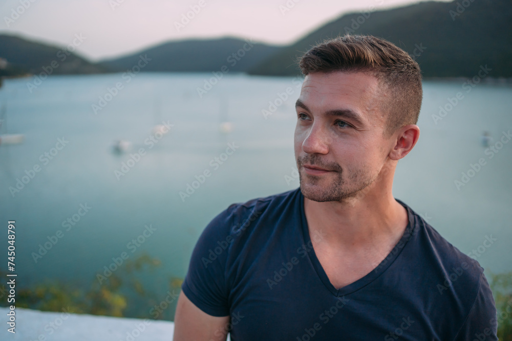 Fototapeta premium A man in a dark T-shirt on the background of a lake. In the background there are green mountains covered with vegetation. There are boats and yachts floating on the lake