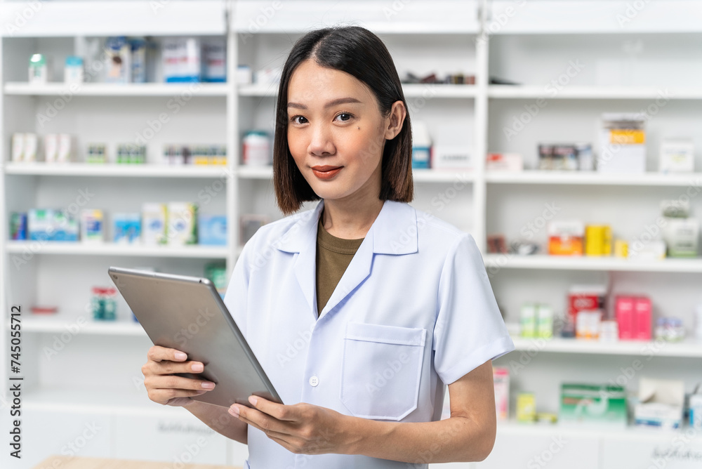 Happy beautiful asian female pharmacist wearing lab coat standing and holding tablet, She looking at camera and feels good, trustworthy and proud of his work in the pharmacy drugstore.