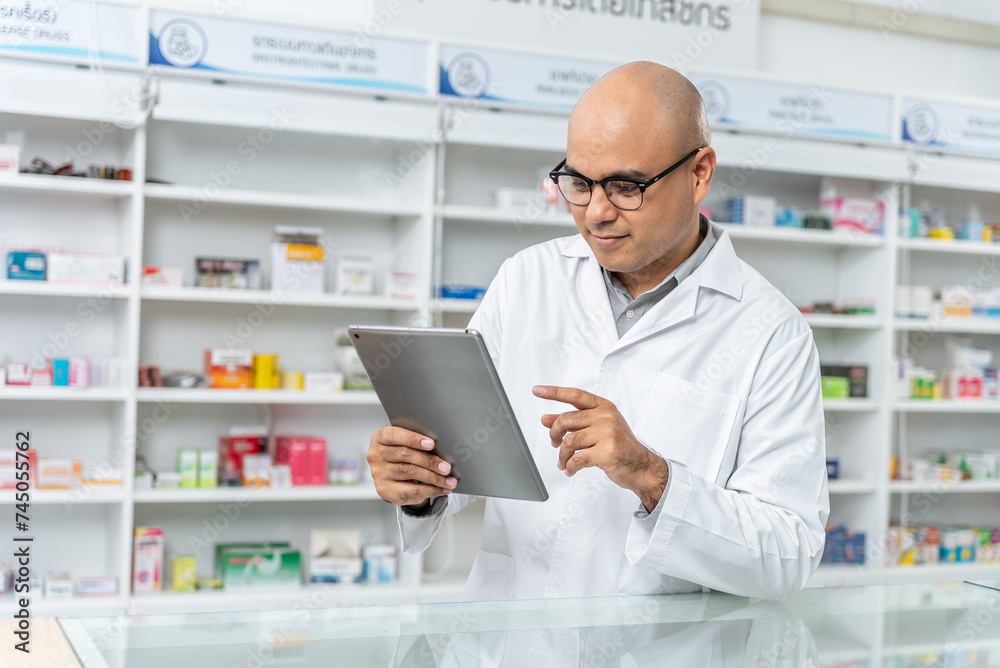 Happy handsome asian male pharmacist wearing eyeglasses and lab coat working with tablet. He feels good, trustworthy and proud of his work in the pharmacy drugstore.